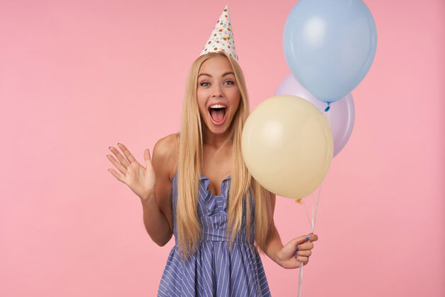五彩喜出望外的年轻漂亮的长发女人 金发在五颜六色的气球里摆姿势 穿着蓝色的夏装 戴着生日帽 在庆祝节日的时候被客人逗乐娱乐姿势圆锥形