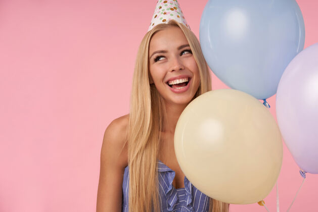 休闲特写镜头中迷人的年轻金发女性一边看一边 面带微笑 在蓝色夏装和生日帽的五彩缤纷的气球里摆姿势 孤立在粉色背景下庆祝裙子气球