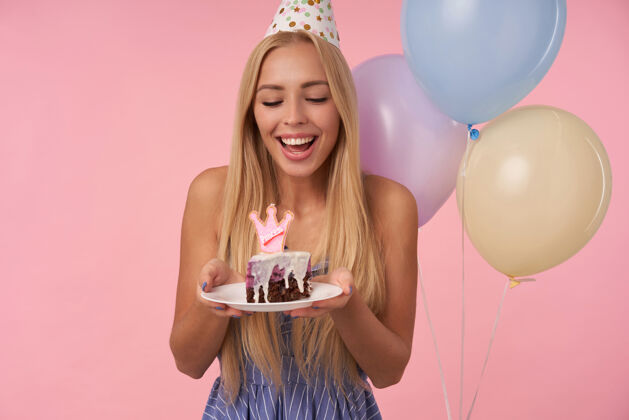 20多岁身着蓝色夏装庆祝节日的快乐的年轻长发女人的肖像 与生日蛋糕一起在五颜六色的气球中摆姿势 孤立在粉色背景上庆祝女士吹