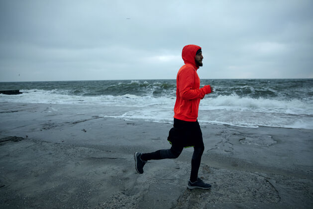 帅哥年轻活跃的胡须男在海边晨练 在寒冷阴沉的天气里沿着海滨奔跑 穿着暖和的运动服健身男模年轻黑暗黑发