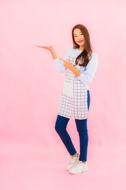 新鲜肖像美丽的亚洲年轻女子在厨房穿着围裙在粉红色的隔离墙上工人烹饪美食