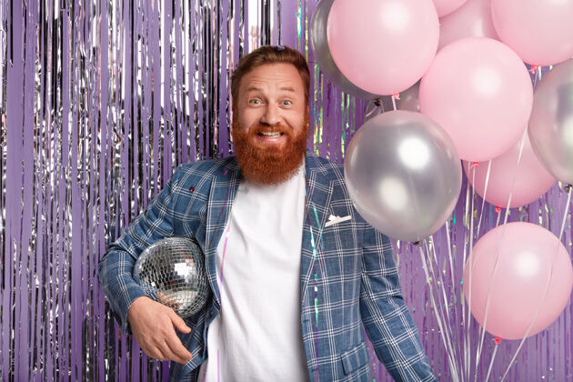 夹克快乐的帅哥在工作中升职后举办派对 手拿迪斯科舞会和一堆气球 开心地笑着 穿着优雅的夹克 站在紫色的金属丝窗帘前 享受着响亮的音乐风格服装外套