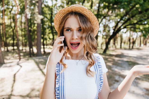 成人震惊的金发女孩站在公园里讲电话户外拍摄的戴着帽子的美丽年轻女子在交谈中表示惊讶快乐树人