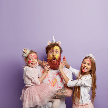 绘画惊艳红发男子戴独角兽角 和两个小女孩玩耍 玩乐色彩 画脸蛋和衣服 心情好 隔着紫色的墙家庭观念感觉积极父亲