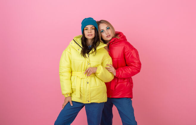 秋季两位迷人的时尚女性在粉色背景上合影 身着红黄相间的缤纷冬季羽绒服 暖衣时尚潮流户外衣服女性