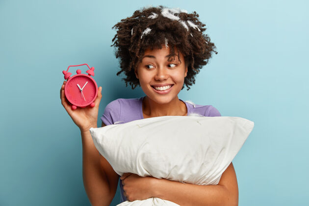 年轻可爱的非洲裔美国女人的照片 卷发 闹钟上显示时间 很高兴晚上睡得很长 微笑积极 抱着柔软的白色枕头 隔着蓝色的墙壁睡觉睡眠开放风格