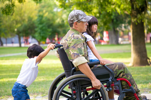 军队残疾退伍军人和两个孩子在公园散步女孩坐在爸爸的腿上 男孩推着轮椅退伍军人或残疾概念轮椅侧孩子