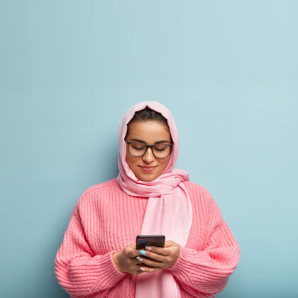 小玩意戴着头巾的迷人友好的女人 追随伊斯兰教 关注明天的日程安排 使用现代手机 在互联网上搜索信息 穿着超大套头衫 隔离在蓝色的墙上伊斯兰手机女人