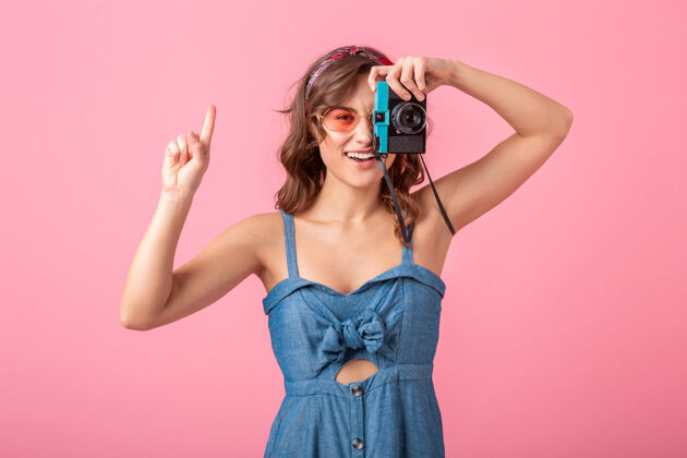 带着迷人的微笑女人在老式相机上拍照 手指向上 穿着牛仔裙 戴着粉色背景的太阳镜连衣裙情感复古