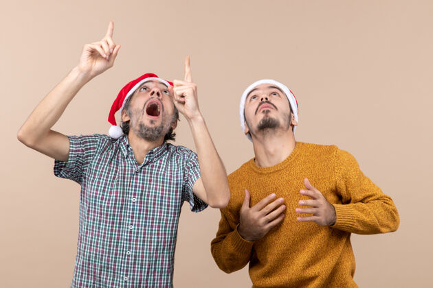 伙计们正面图：两个戴着圣诞帽的男人 一个在米色背景下向另一个展示着什么脸男性两个惊奇的家伙