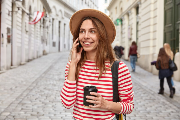 条纹美丽的微笑游客漫步在城市 有电话交谈 举行外卖咖啡 集中到远处的某个地方使用饮用牙关