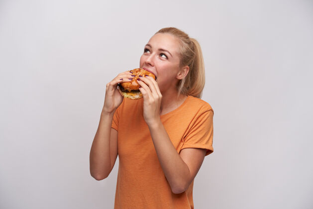 汉堡一张室内照片 一位年轻的金发碧眼的女士 梳着马尾辫 穿着休闲服 一边在白色背景下摆姿势 一边吃着美味的汉堡包 一边积极地看着一边食物马尾辫快餐