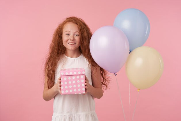 乐趣摄影棚拍摄的卷发女孩儿 长着狐狸精的头发 手里拿着礼物包装盒 兴奋又惊讶地收到生日礼物 开心地看着粉色背景下的相机节日高兴孩子