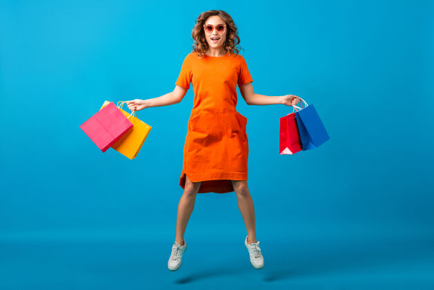 休闲迷人的快乐微笑时尚女性购物狂穿着橙色时髦的超大号连衣裙蹦蹦跳跳拿着蓝色背景的购物袋女孩购物者零售