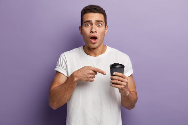 震惊镜头中的年轻人目瞪口呆地指着外卖咖啡 惊讶地发现空纸杯里没有任何饮料 穿着白色休闲衬衫 张嘴 与紫色的墙壁隔绝广告咖啡广告