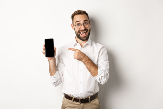 员工兴奋的帅哥拿出手机 手指着屏幕微笑着 站在白色的背景下男人复制应用程序
