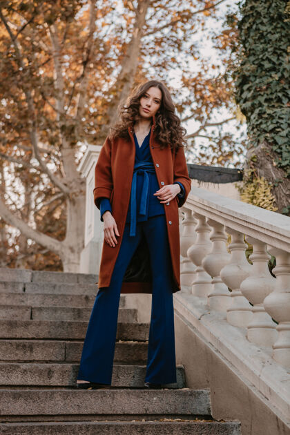 穿着美丽时尚的微笑瘦身女子卷发走在街头楼梯上穿着温暖的棕色外套和蓝色西装 秋季时尚街头风外套秋天优雅