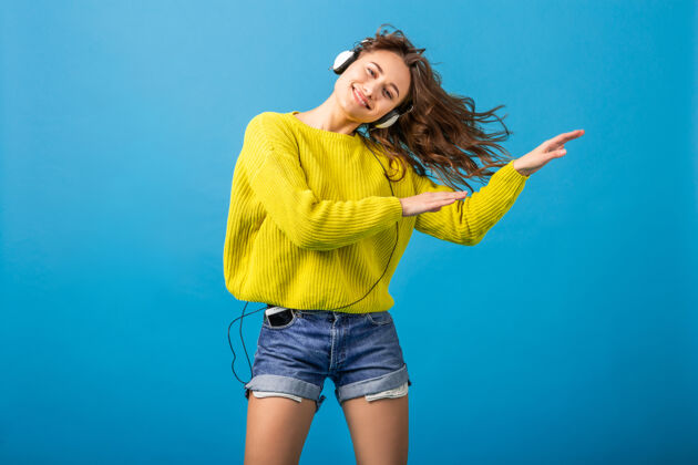 蓝色迷人的笑容可掬的快乐女人戴着耳机跳舞听音乐 穿着时髦的服装 隔离在蓝色的工作室背景下 穿着短裤和黄色毛衣情感跳跃耳机