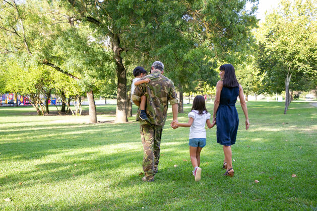 军队一家人在公园的草地上散步 父亲穿着迷彩服 抱着儿子 和妻子孩子一起享受周末 家庭团聚 回家的理念团聚散步年轻