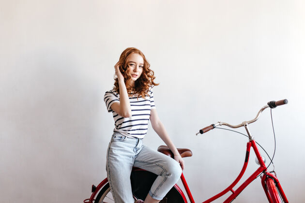 头发穿着牛仔裤坐在自行车上的优雅女士的室内肖像可爱的姜黄色女士与自行车合影自行车手女性波浪