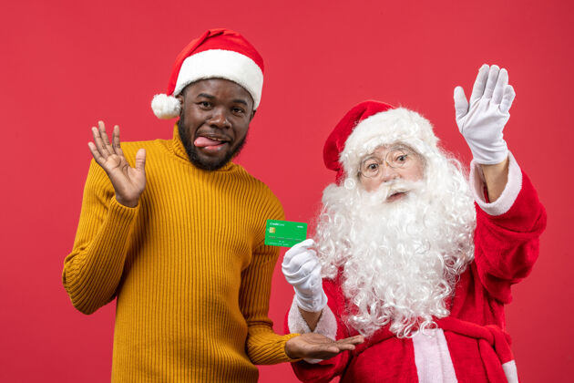 圣诞老人红墙上的圣诞老人和拿着绿色银行卡的年轻人的正视图男性圣诞快乐圣诞