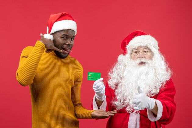 年轻红墙上的圣诞老人和拿着绿色银行卡的年轻人的正视图人们节日前面