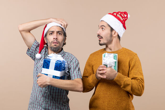 两个正面图两个戴着圣诞帽拿着圣诞礼物的困惑的家伙 一个在米色孤立的背景上挠头帽子圣诞节家伙