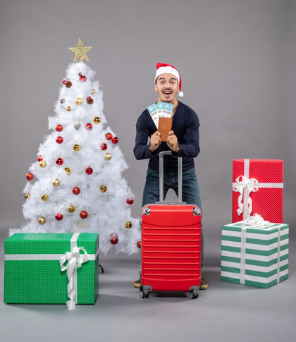礼物快乐的年轻人拿着红色的手提箱 在灰色的墙上展示他的旅行票盒子旅行圣诞老人