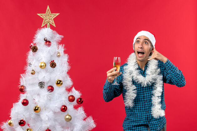 圣诞老人圣诞心情快乐的年轻人 戴着圣诞老人帽 穿着蓝色条纹衬衫 举起一杯葡萄酒 在圣诞树旁做着“打电话给我”的手势玻璃年庆祝