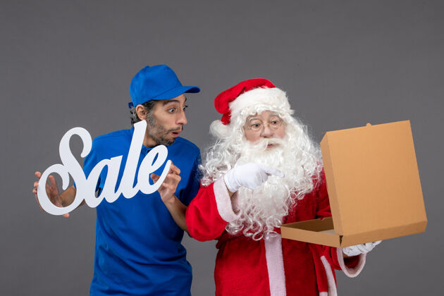 男圣诞老人的正面图 男性信使手持销售横幅 灰色墙上有食品盒十二月圣诞快乐盒子