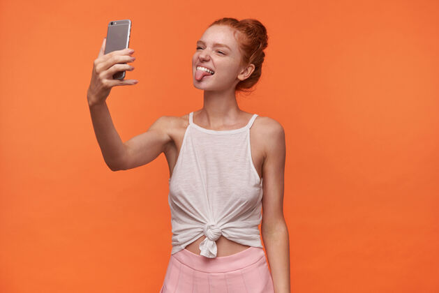智能手机照片中 迷人的年轻女士扎着一头狐狸精的头发 手里拿着智能手机 一脸滑稽地看着相机 对着相机傻笑 露出舌头 孤立在橙色背景下快乐休闲红发