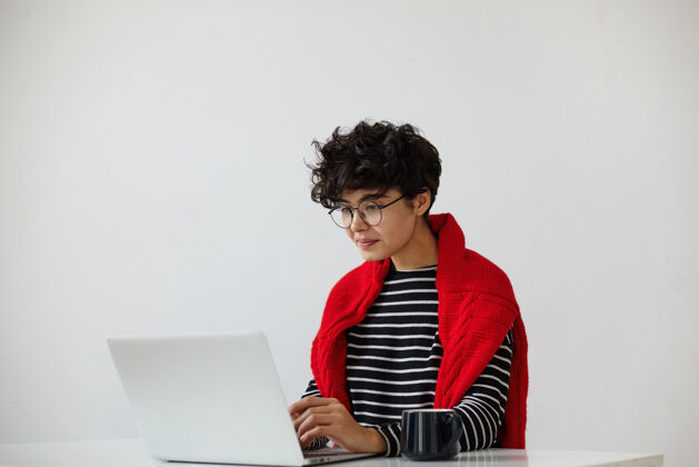时尚年轻漂亮的黑发卷发女士 留着短发 穿着时髦的休闲服 坐在白色背景下的桌子旁 拿着笔记本电脑针织室内杯子
