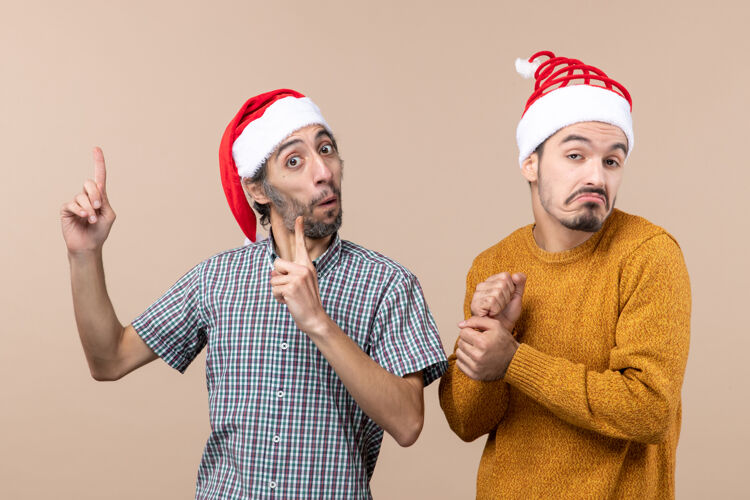 观点前视图两个戴着圣诞帽的困惑的家伙一个手指在米色孤立的背景上展示着什么展示微笑成人
