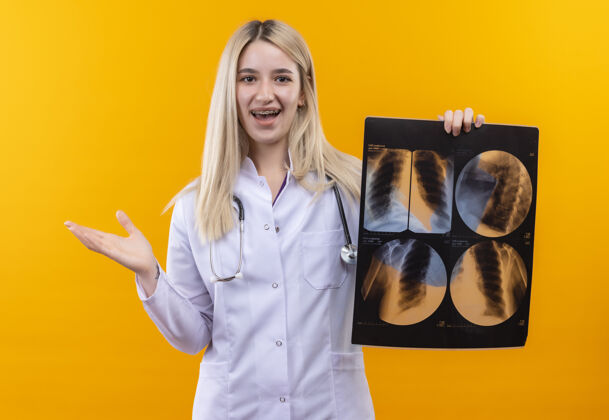 牙科微笑的医生年轻的女孩穿着医用长袍戴着听诊器和牙套拿着x射线指向隔离黄色背景的一侧听诊器X光侧