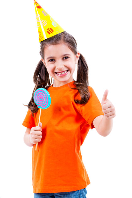 五颜六色身着橙色t恤和派对帽的可爱小女孩的肖像 彩色糖果显示竖起大拇指的手势-白色隔离糖果竖起大拇指食物