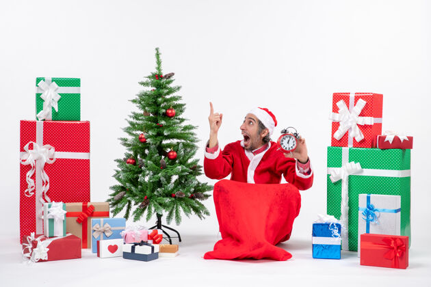 惊讶惊讶的年轻人坐在地上庆祝新年或圣诞节 拿着礼物和白色背景装饰圣诞树附近的时钟男人抱着礼物