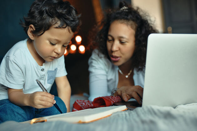 人专注的黑皮肤小男孩学习字母表 写在抄写本上的字母 坐在床上和他的年轻母亲使用便携式电脑远程工作远程房间工作