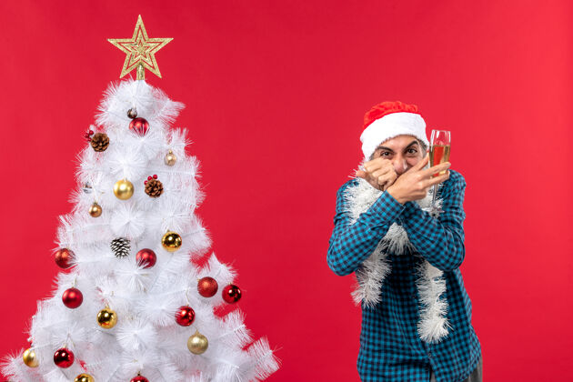 疯狂圣诞心情快乐疯狂情绪化的年轻人戴着圣诞老人的帽子 穿着蓝色条纹衬衫 在圣诞树旁举起一杯葡萄酒提高年轻白色