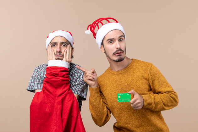 两个前视图两个家伙一个穿着圣诞外套 手放在脸上 另一个拿着信用卡 背景是米色的服装手伙计们