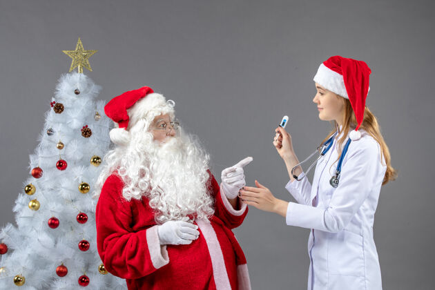 快乐圣诞老人和女医生的前视图 女医生拿着测温仪在灰色的墙上服装人温度