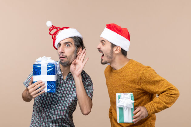 成人正面图两个好奇的家伙闲聊和持有米黄色孤立背景的圣诞礼物米色帽子抱着