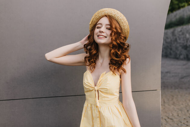 时尚穿着复古长裙的苗条浪漫女孩在灰色墙上欢笑戴着帽子的高加索姜黄色女模特的照片有趣欢快愉悦