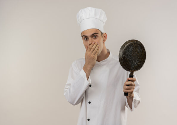 嘴穿着厨师制服的年轻男厨师惊讶地用手捂住嘴 拿着有复印空间的煎锅惊讶厨师戴着