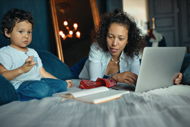 小可爱的混血小男孩坐在床上画画 而他的年轻母亲使用便携式电脑进行远程工作的室内肖像拉丁语玩耍小