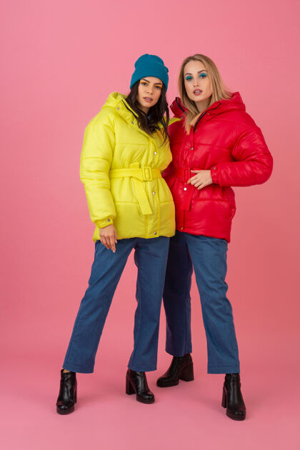保暖两个迷人的女孩在粉红色的背景下 穿着鲜艳的红色和黄色的彩色冬季羽绒服摆姿势保暖衣服女性时尚