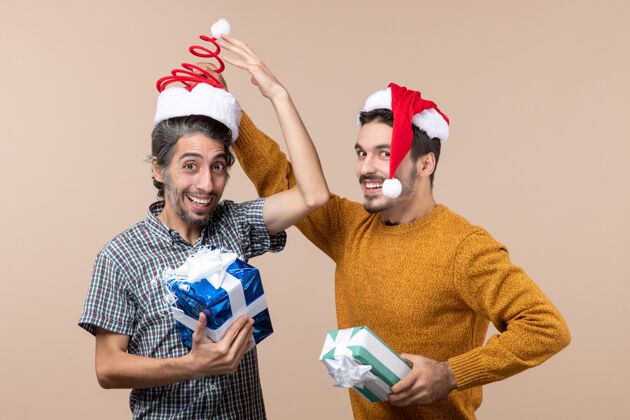 米色正面图：两个快乐的家伙拿着圣诞礼物 一个正在摘下另一个的帽子 背景是米色的圣诞节男性成人