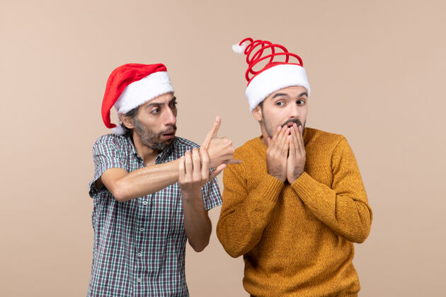 微笑前视图两个戴着圣诞帽的困惑的家伙一个在米色孤立背景上向另一个展示标志肖像男性视图