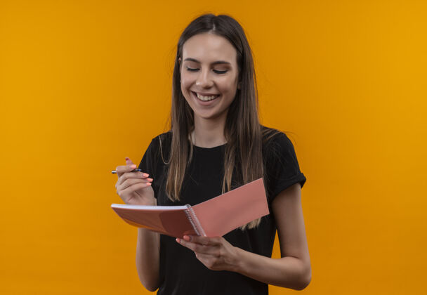 微笑微笑着的年轻白人女孩穿着黑色t恤 在孤立的橙色背景下看着手里的笔记本橙色黑色看