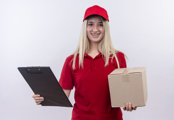 帽子微笑的小女孩穿着红色t恤 戴着帽子 戴着牙套 拿着盒子和剪贴板 背景是白色的白色交货微笑