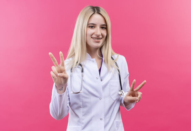 长袍微笑的医生年轻的金发女孩穿着医用长袍戴着听诊器和牙套 在孤立的粉红色背景上用双手做着和平的手势显示女孩两者
